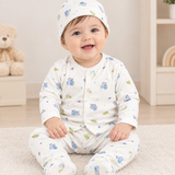 A smiling baby sitting on a white carpet wearing a matching footed sleeper outfit and bonnet with a blue and green cartoon animal print on a white background.