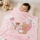 A baby sleeping peacefully under a pink plush blanket with cartoon teddy bears, a crescent moon, and the text 'ALL OF ME LOVES ALL OF YOU'. A beige teddy bear sits on a wooden shelf in the background.