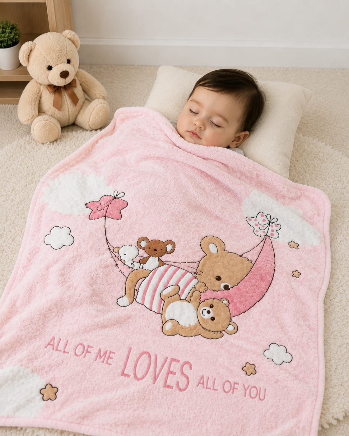 A baby sleeping peacefully under a pink plush blanket with cartoon teddy bears, a crescent moon, and the text 'ALL OF ME LOVES ALL OF YOU'. A beige teddy bear sits on a wooden shelf in the background.