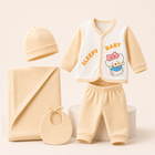 A baby clothing set arranged on a light beige studio background, including a cream and pale yellow long-sleeve vest with 'SLEEPY BABY' text and a cartoon bear graphic, matching pale yellow pants, a cream beanie, a folded cream blanket, and a cream bib.