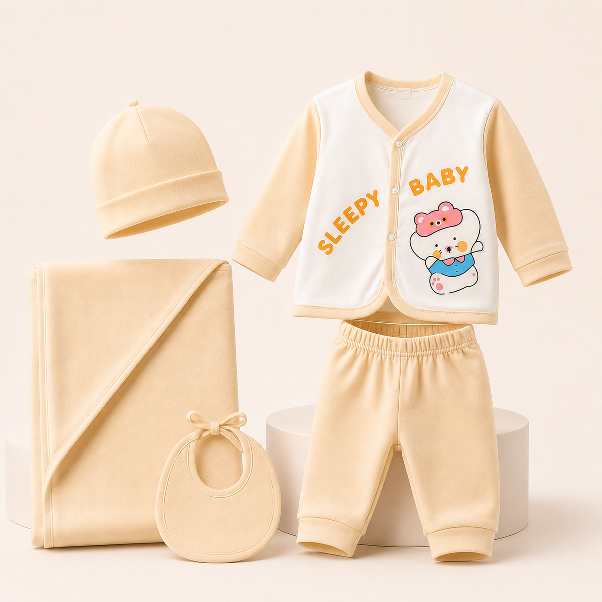 A baby clothing set arranged on a light beige studio background, including a cream and pale yellow long-sleeve vest with 'SLEEPY BABY' text and a cartoon bear graphic, matching pale yellow pants, a cream beanie, a folded cream blanket, and a cream bib.