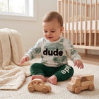 A baby sitting on a cream rug in a nursery, wearing a white long-sleeve shirt with a green tie-dye pattern and the word 'dude' printed in black across the chest, paired with dark green pants. The baby is playing with wooden toys, with a crib and a window in the background.